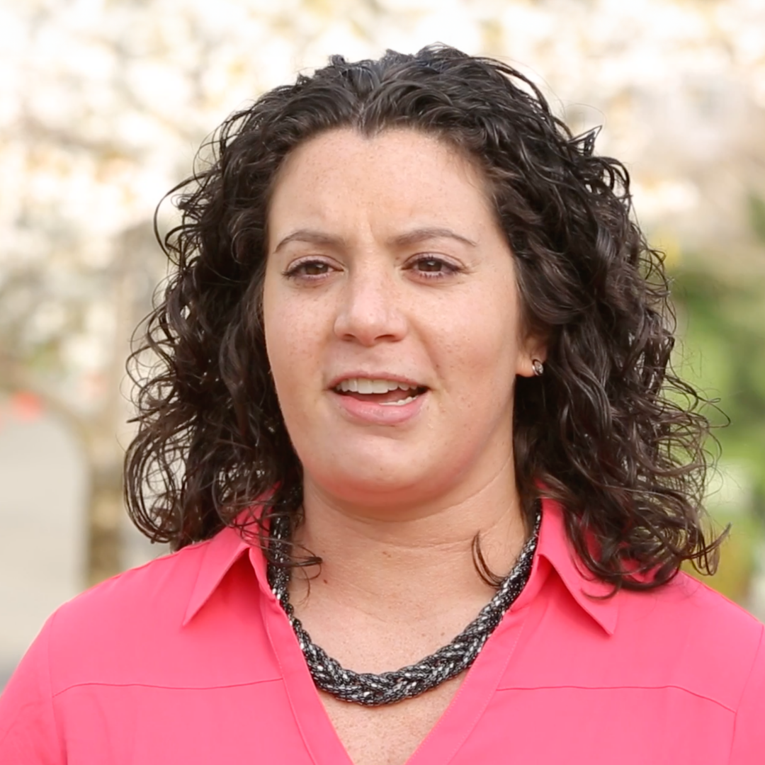 A woman with curly dark hair, wearing a bright pink blouse and a black beaded necklace, stands outdoors with blurred trees and greenery in the background.